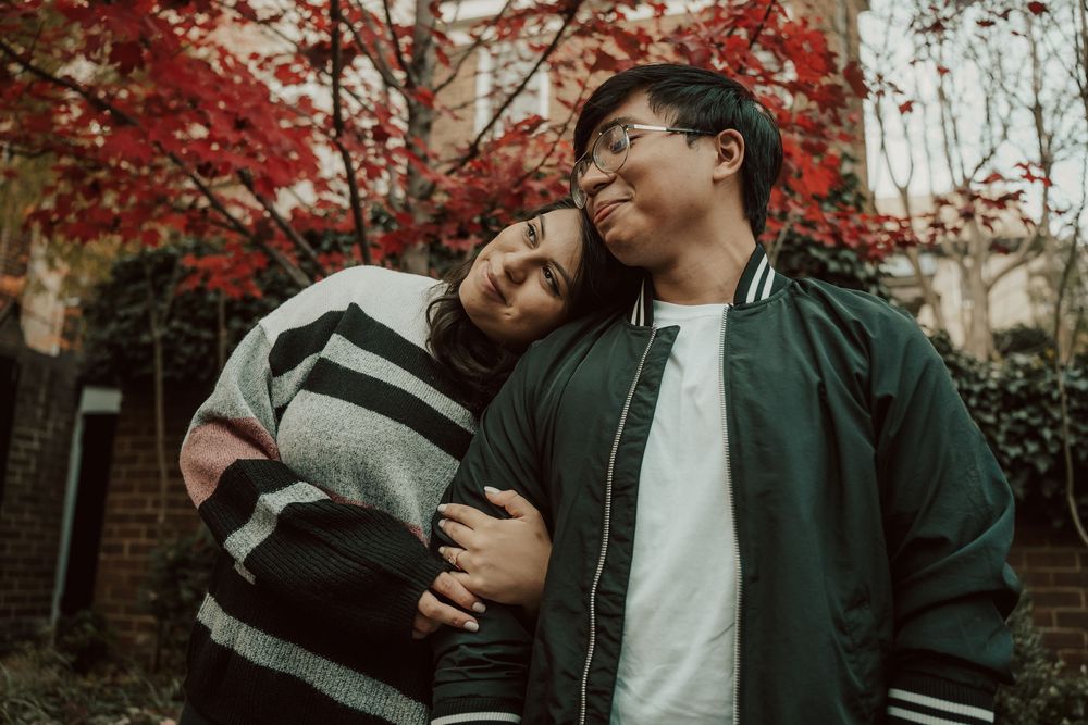 A couple in dark clothing embraces tenderly against a backdrop of autumn foliage and brick walls in Old town Alexandria